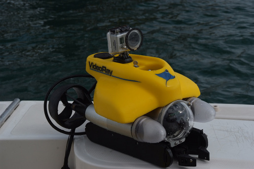 A remotely operated vehicle (ROV) resting on a boat with the ocean in the background. The ROV is a small, motorized device with an action camera mounted on top.